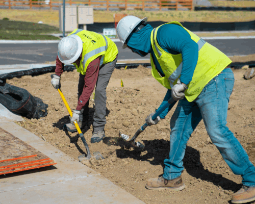 Two construction workers dig by a sidewalk at a construction site.