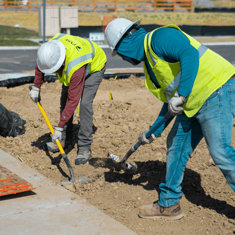 Two construction workers dig by a sidewalk at a construction site.